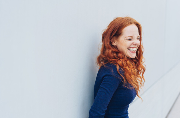 Woman with red hair in a blue dress standing against a wall and smiling at United Smile Centres in Louisville, KY